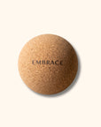 A round cork massage ball with the word 'EMBRACE' printed in black text on its surface, set against a solid cream-colored background.